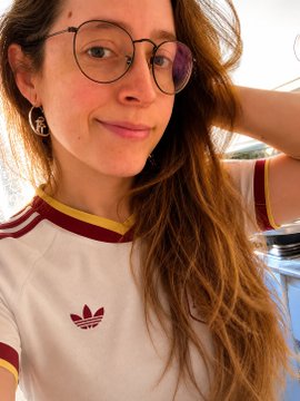 A young woman with long brown hair and round glasses wears a cream-colored Adidas t-shirt with maroon and yellow striped collar, smiling softly in a selfie. The casual travel photo complements the caption's mention of visiting Portugal, while the Adidas branding ties in with the promotional mention of BCgame.