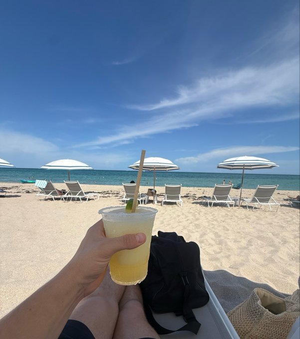 A person's hand holds a frozen yellow drink on a sandy beach, with beach chairs and striped umbrellas overlooking the ocean under a blue sky.