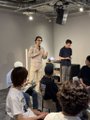 A small image showing two men standing at the front of a classroom or lecture hall, addressing an audience of seated attendees.
