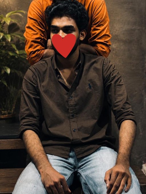 A young man in a brown button-up shirt sitting down, with a large red heart emoji covering his face, while another person in an orange shirt stands behind him.