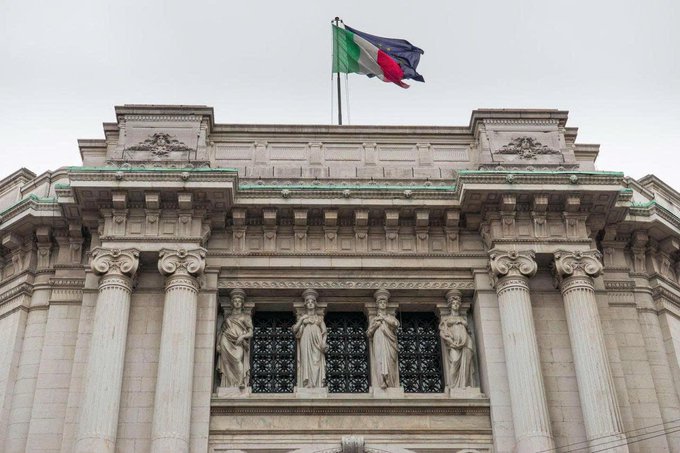 An architectural photograph of a grand neoclassical building featuring four caryatid statues between Ionic columns, with a green flag flying above the roofline against an overcast sky.