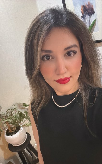 A selfie of a woman with long dark hair, red lipstick, and a pearl necklace, wearing a black top next to a potted plant.