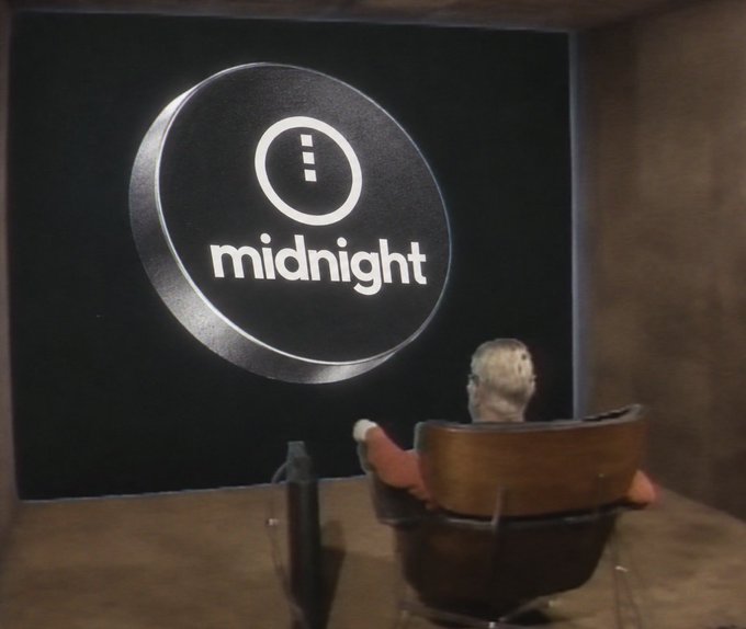 A small figurine of a person sits in a chair in front of a glowing circular sign that reads "midnight" with an information icon.