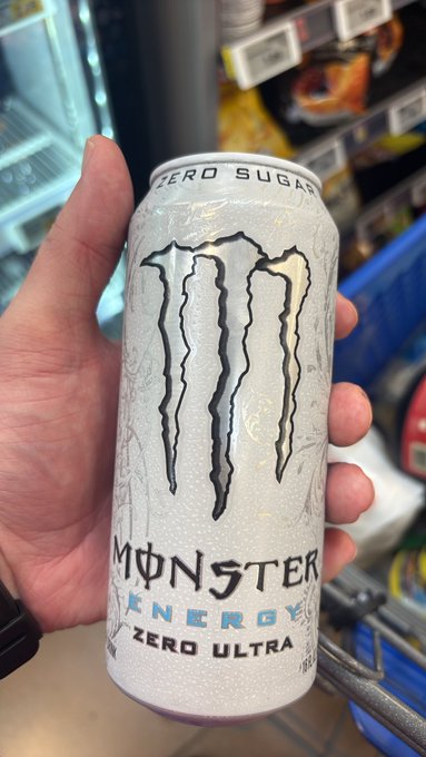 A hand holds up a white, condensation-covered can of 'Monster Energy Zero Ultra' (Zero Sugar) in front of a refrigerated store display case.