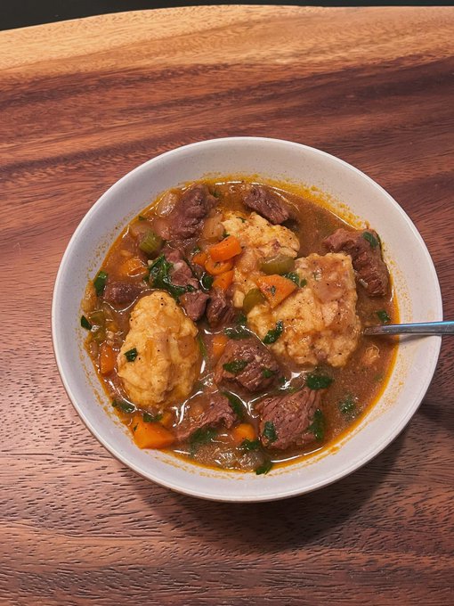 A cozy white bowl filled with hearty beef and vegetable stew, featuring tender chunks of meat, carrots, and large golden dumplings, sits on a warm wooden table. The homemade-looking dish perfectly embodies the comforting, home-cooked vibe of the caption.