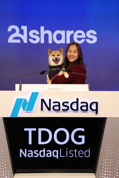 A woman holds a Shiba Inu dog while standing at a Nasdaq podium, with '21shares' on the backdrop and a screen displaying 'TDOG NasdaqListed'.
