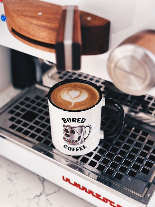 A cappuccino with heart latte art in a white mug that reads 'BORED COFFEE' sitting on the drip tray of a La Marzocco espresso machine.