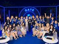 A large group of people poses for a photo on stage in front of a backdrop reading 'GATE', with many individuals raising their hands in celebration.