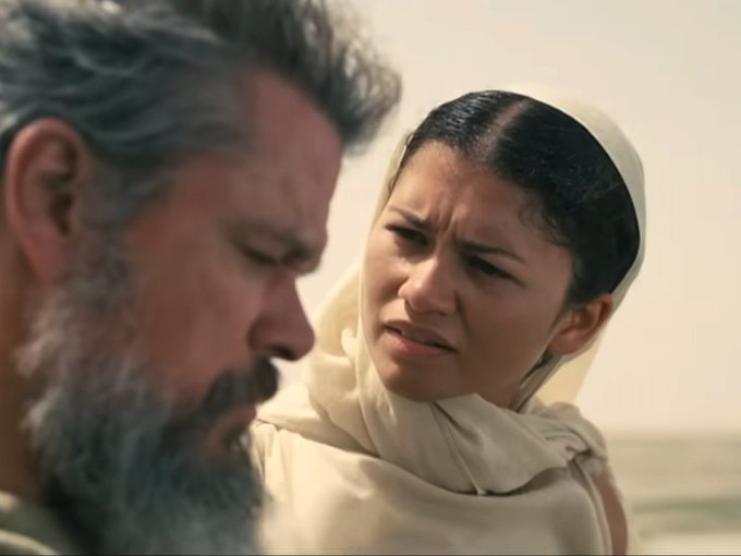 A cinematic close-up of an older man with a gray beard alongside a woman wearing a white headscarf, both with serious expressions.