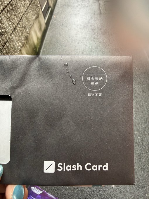 A black envelope labeled 'Slash Card' with Japanese text '料金後納郵便' (postage paid) and '転送不要' (do not forward) stamp, held in front of a paved surface.
