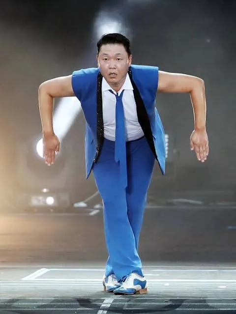 A man in a blue suit with a white shirt and blue tie strikes an energetic pose, leaning forward with arms outstretched on a stage with dramatic lighting.