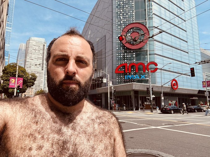 A shirtless bearded man taking a selfie in front of an AMC IMAX theater located in a busy city area with tall buildings.