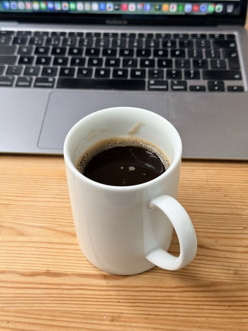 A white ceramic mug filled with black coffee sits on a wooden desk in front of a MacBook Pro, with the visible apps including Safari, Mail, and Notes in the menu bar—suggesting the coffee drinker is fueling up for a turbulent market week.