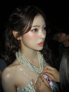Photo of a young woman with dark hair in pigtails wearing layered pearl necklaces and a sparkly outfit, looking to the side in a dimly lit setting.