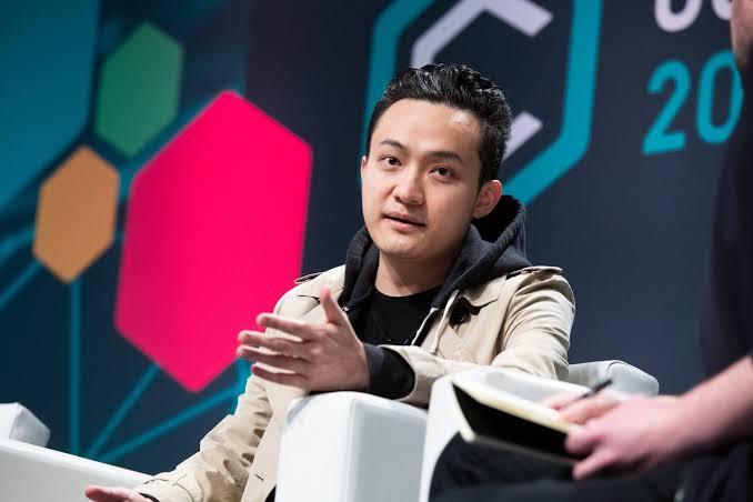 An Asian man in a tan jacket and black hoodie speaks while gesturing with his hand at what appears to be a conference or panel, with a colorful hexagonal backdrop partially visible.