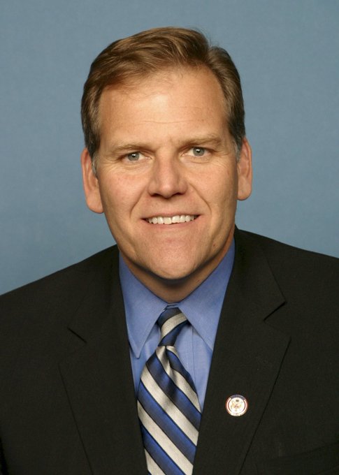 A professional headshot of a smiling middle-aged man with short brown hair, wearing a black suit, blue shirt, and blue-and-black striped tie, featuring a small lapel pin. The image relates to the post as it appears to be an official portrait of Mike Rogers, who won Michigan's Republican Senate primary with Trump's endorsement.