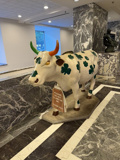 A decorative cow statue with green clover spots and colorful horns stands on a marble floor, displaying a sign that reads 'PLEASE DO NOT CLIMB ON ME THANK YOU!'.