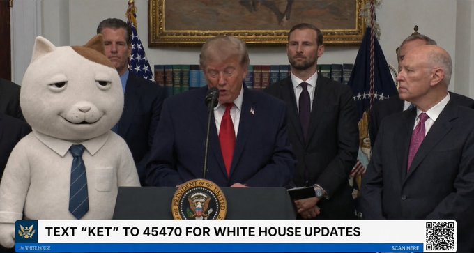 A screenshot from a White House press briefing showing President Donald Trump at the podium speaking, with a digitally edited cat mascot in a white shirt and blue tie standing to his left, and a banner reading 'TEXT "KET" TO 45470 FOR WHITE HOUSE UPDATES'.