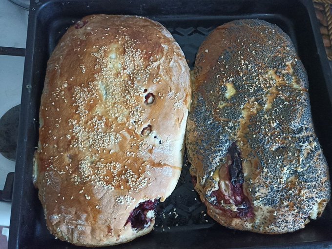 Two freshly baked loaves of bread with fruit filling in a black baking tray; one is topped with sesame seeds and the other with poppy seeds.
