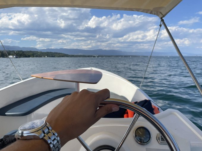 A hand wearing a silver watch gripping the steering wheel of a boat on open water with mountains and clouds in the distance.