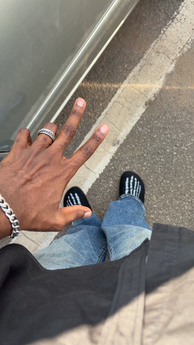 A first-person perspective looking down at a person's hand making a gesture, wearing a silver ring and chain bracelet, with black sneakers and blue jeans visible on a sidewalk.