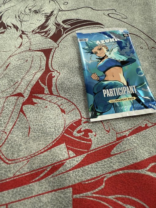 An 'AZUKI' trading card game Participant Tournament Pack booster pack resting on a gray and red illustrated playmat featuring an anime-style character.