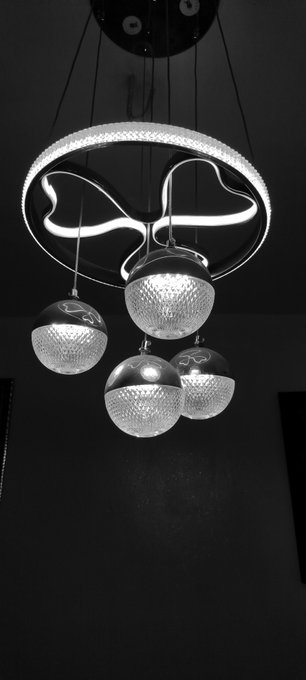 A modern pendant light fixture features a circular ring with a wavy interior tube and four textured crystal globe pendants hanging below, photographed in black and white.