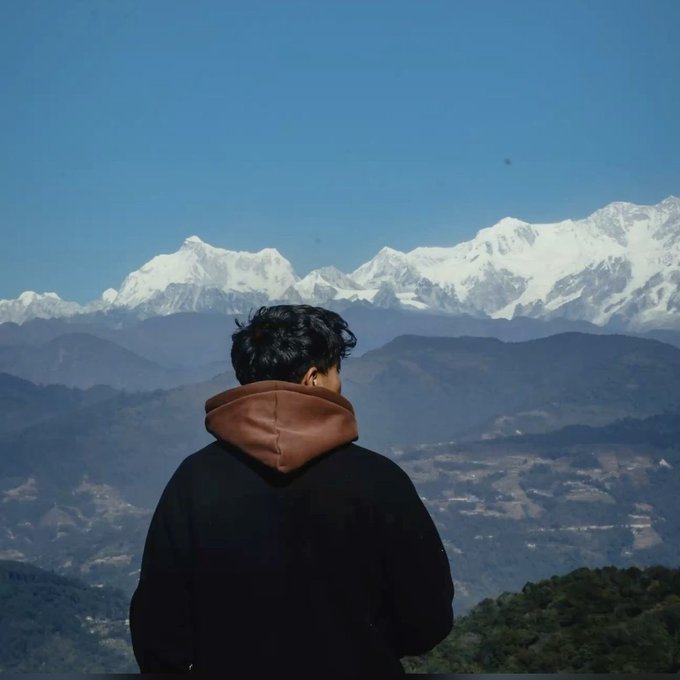 A photograph of a man from behind, wearing a black hoodie with brown trim, gazing out at a stunning panoramic view of snow-capped Himalayan mountains under a clear blue sky.