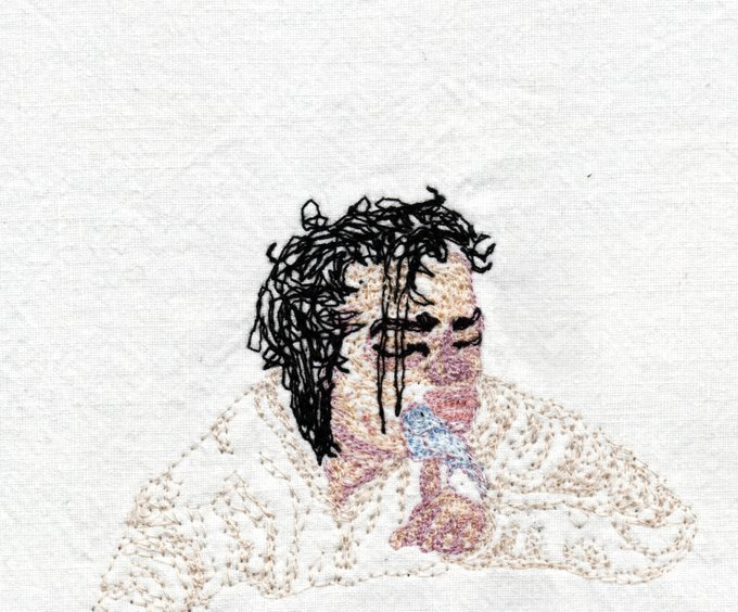 An embroidered textile artwork on white fabric depicting a portrait of a person with dark, scribbled hair and muted pink, blue, and beige tones on their clothing.