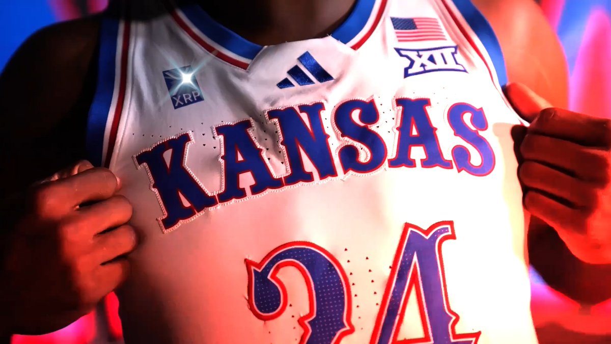 A close-up of a Kansas Jayhawks basketball jersey featuring the Big 12 (XII) logo, American flag patch, Adidas logo, and an XRP sponsor patch, with the number 34 visible.