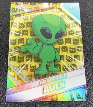 A holographic trading card titled 'ADAPTABLE ALIEN' featuring a green alien character with a peace sign gesture on a patterned yellow background.