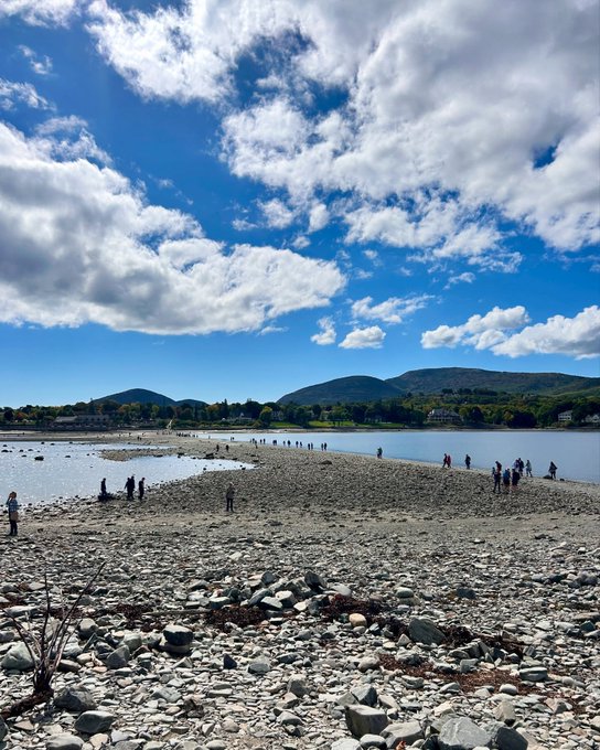 A wide landscape view shows people walking along a rocky riverbed or sandbar beside a calm river, with forested hills in the distance under a blue sky with scattered white clouds.