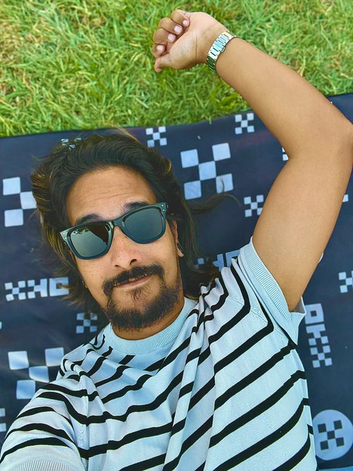 A bearded man in sunglasses and a striped shirt lies on a pixel-patterned blanket in the grass, one arm raised, playfully taking the "touch grass" advice literally.