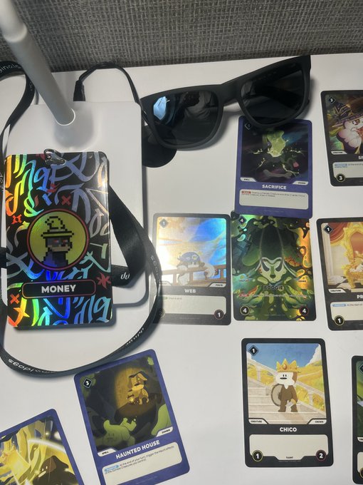 A display of collectible NFT trading cards featuring characters like Money, Wee, Sacrifice, and Chico, alongside a 'Haunted House' card, with black sunglasses and a lanyard resting on the table.