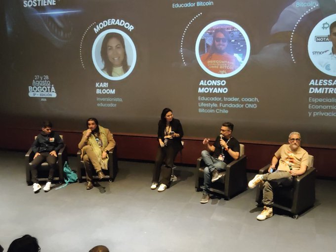 A panel discussion on a stage with five seated speakers, with a large screen behind them displaying headshots and names including moderator Kari Bloom, Alonso Moyano, and Aless(andra) Dmitru; the event is branded as "BOGOTA 9° EDICION" with dates "27 y 28 Agosto."