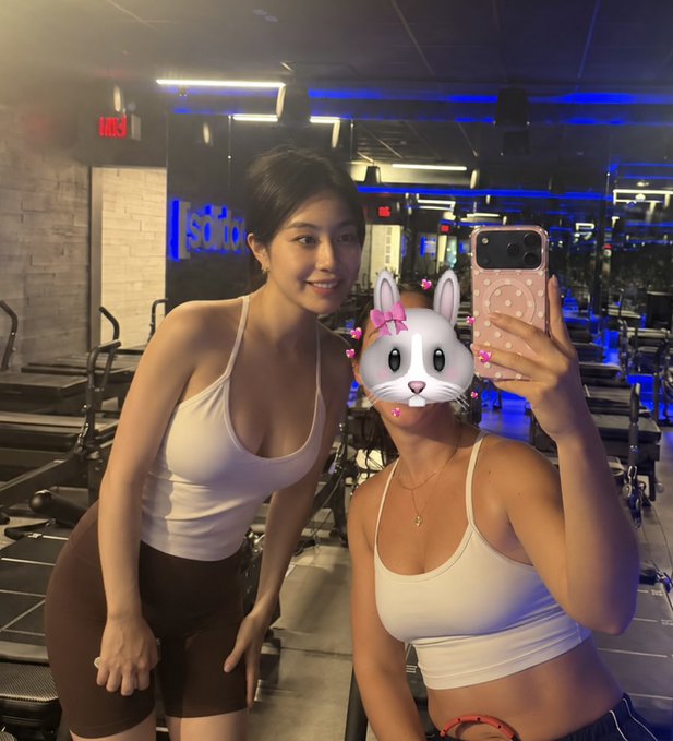 Two women in white tops and dark shorts pose for a mirror selfie inside a gym with blue lighting, with one woman's face covered by a bunny filter.