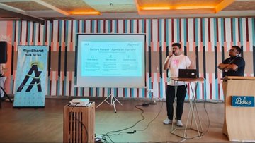The image shows a presentation taking place at an event, with two men standing behind a podium giving a speech. The room has a large screen displaying information related to battery technology, specifically focusing on the Algorand India Dev Retreat. The speakers are discussing the importance of battery powered EV batteries and their role in the industry. 

There is also a laptop placed near one of the speakers, possibly used for visual aids or data sharing during the presentation. A few other people can be seen in the background, likely attending the event as well.