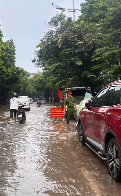 A flooded street scene with people wading through water, a police officer in green directing traffic, and a red car parked on the side.