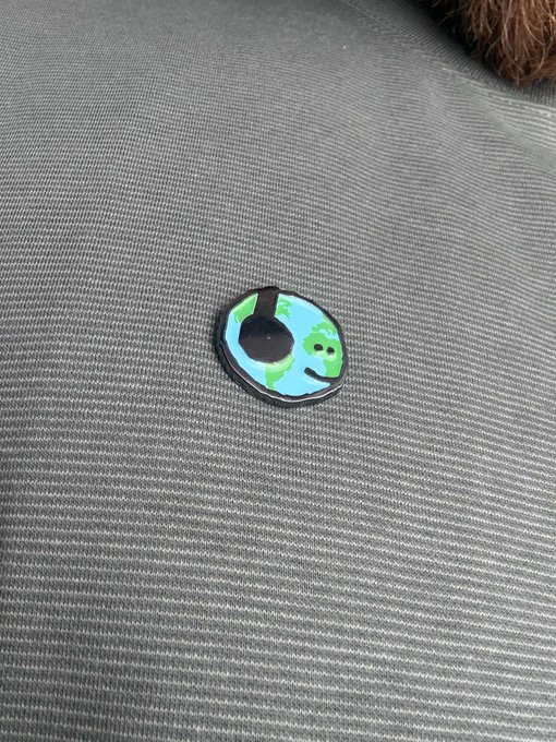 A small circular pin featuring a green cartoon character (resembling a Shiba Inu meme) sits on a gray fabric car seat, fitting the casual "gm mfers" greeting with its playful, meme-inspired crypto culture vibe.