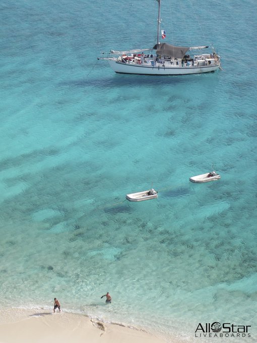 Aerial view of a turquoise sea with a sailboat anchored in clear water, two small dinghies floating nearby, and people on the sandy beach below; watermark reads 'All Star Liveaboards'.