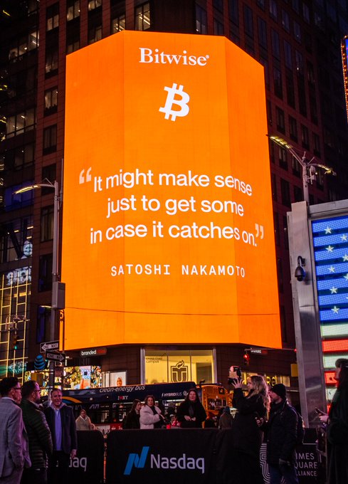 A large orange billboard in Times Square displays the Bitwise logo and Bitcoin symbol alongside a quote from Satoshi Nakamoto: "It might make sense just to get some in case it catches on," with the Nasdaq logo visible at the base. The image supports the post's message about Bitcoin's growing prominence, showcasing how the cryptocurrency is being prominently featured on major advertising displays in one of the world's most iconic financial hubs.