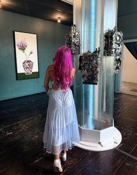 Person with long magenta hair in a blue dress viewing art pieces displayed in a gallery.