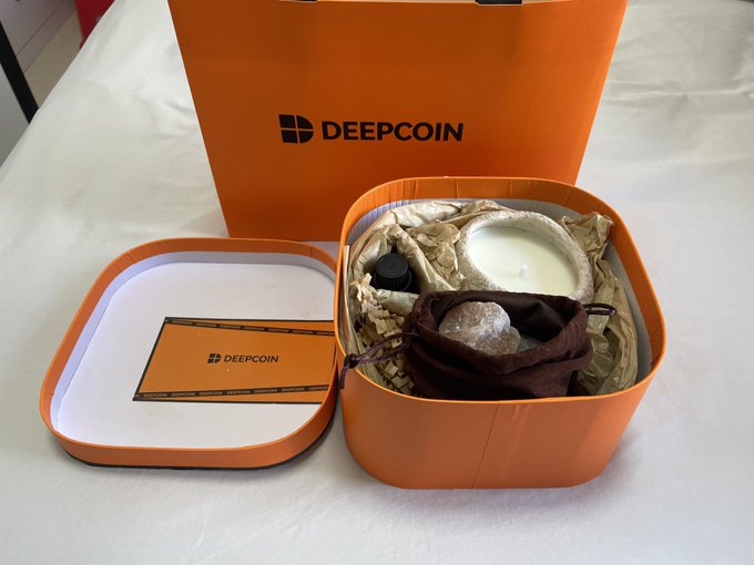 An orange DEEPCOIN branded gift box opened to reveal its contents including a small bottle, a pouch with a wrapped item, and a candle or container with a white substance, alongside a DEEPCOIN branded card.