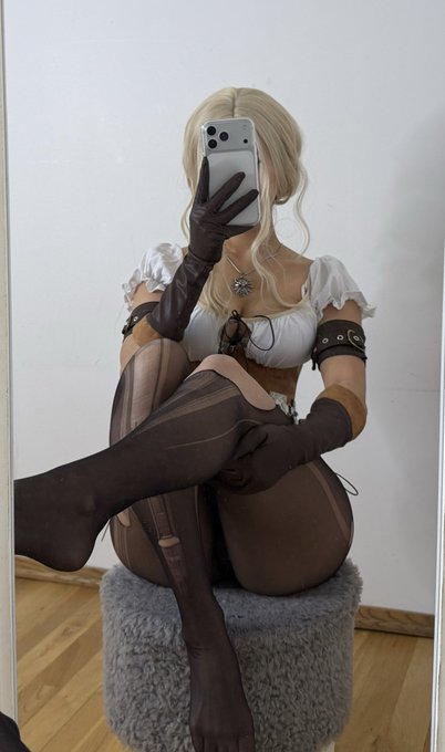 A person dressed in a cosplay outfit takes a mirror selfie with an iPhone, wearing long brown gloves and stockings while seated on a fluffy stool. The blonde wig, white top with puffed sleeves, and the medallion necklace resemble the signature look of Ciri from The Witcher series.