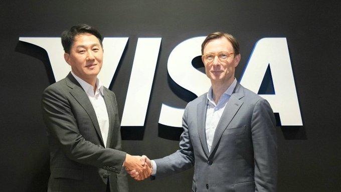 Two men in business suits shake hands while standing in front of a large "VISA" logo on a dark wall, illustrating the partnership announcement between Visa and South Korea's Dunamu to explore stablecoin payments and AI-powered financial services.