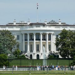 The White House, with an American flag flying atop its neoclassical South Portico, stands behind a perimeter fence as tourists gather on the sidewalk. The image visually anchors the breaking news about the Trump administration's push for Congress to pass the CLARITY Act, reinforcing the official source of the statement.