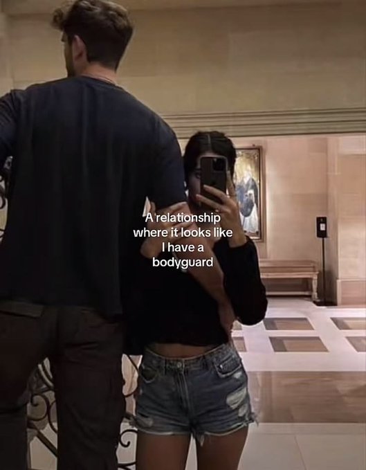 A mirror selfie with text overlay reading "A relationship where it looks like I have a bodyguard", showing a man standing protectively next to the woman taking the photo.
