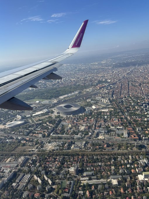 Aerial view from a Wizz Air airplane window showing a wing and a cityscape below with stadiums, dense housing, and streets.