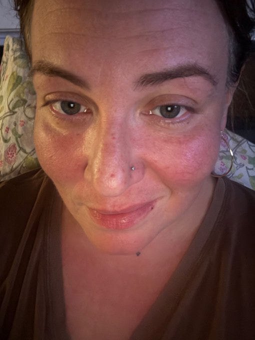 A close-up selfie of a woman with flushed skin and a nose piercing, sitting against a floral patterned pillow.