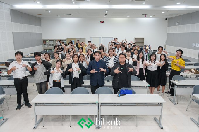 A large group of people pose for a group photo inside a classroom or conference room, making hand gestures together, with a 'Bitkub Academy' logo displayed at the bottom.